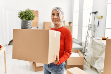 Orta yaşlı, kır saçlı, gülümseyen bir kadın yeni evinde kendine güvenen bir paket tutuyor.