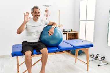 Orta yaşlı İspanyol adam ağrı kesici kliniğinde elinde pilates topu tutarak mutlu ve güler yüzlü hoş geldin jesti diyor. 