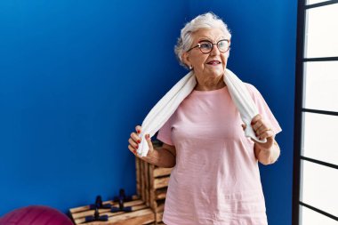 Spor kıyafetleri giyen yaşlı gri saçlı kadın spor merkezinde kendine gülerek duruyor.