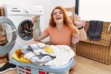 Kirli çamaşırlarını çamaşır makinesine koyan genç kızıl saçlı kadın mutlu ve güler yüzlü bir şekilde selam veriyor. 
