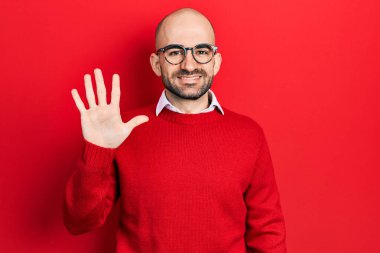 Genç kel adam, günlük kıyafetler ve gözlük takıyor. Beş numaralı parmaklarıyla işaret ediyor ve gülümsüyor. Kendinden emin ve mutlu.. 