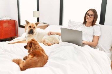 Genç İspanyol kadın, yatak odasında köpeklerle birlikte dizüstü bilgisayar kullanıyor.