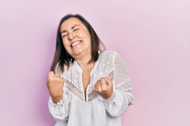 Orta yaşlı İspanyol kadın gündelik giysiler giyip çok mutlu ve heyecanlı bir şekilde kollarını kaldırıp, gülümseyip, başarı için haykırarak kazanan bir jest yapıyor. kutlama konsepti. 