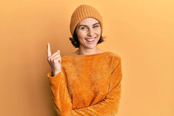 年轻的黑发女人戴着羊毛帽子 穿着冬季毛衣 高兴地微笑着 手指手画脚地指向旁边 — 图库照片