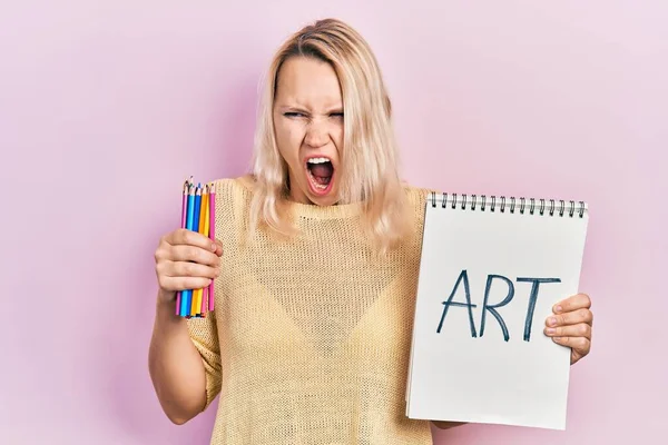 美しい白人ブロンドの女性がアートノートと色鉛筆を持って怒りと怒りで叫んで フラストレーションと激怒叫んで怒っています 怒りと攻撃的な考え方 — ストック写真