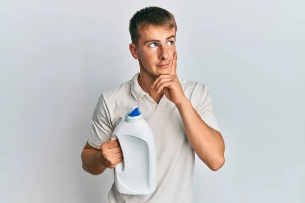 Joven Hombre Caucásico Sosteniendo Botella Detergente Cara Seria Pensando Pregunta —  Fotos de Stock