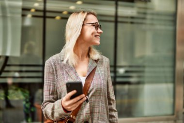 Genç sarışın iş kadını şehirde akıllı telefon kullanarak mutlu bir şekilde gülümsüyor..