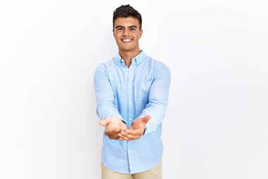 İzole edilmiş arka planda duran İspanyol gömlekli genç adam avuçlarını birbirine uzatarak gülümsüyor ya da el hareketi yapıyor. Bekleme ve koruma 