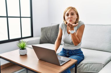 Orta yaşlı sarışın kadın dizüstü bilgisayar kullanıyor ve kameraya bakıyor. Canlı yayında öpüşüyor. Güzel ve seksi. aşk ifadesi. 