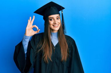 Mezuniyet şapkası ve tören cübbesi giymiş güzel esmer bir kadın gülümsüyor ve el ve parmaklarıyla imza atıyor. başarılı ifade. 