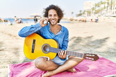 Gitar çalan genç İspanyol adam kumsalda kumların üzerinde oturmuş akıllı telefonla konuşuyor..