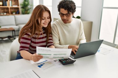 Genç çift evde masada dizüstü bilgisayar kullanarak mutlu bir şekilde gülümsüyor..