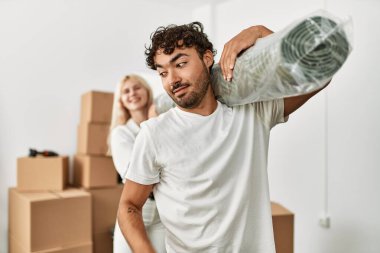 Genç ve güzel bir çift yeni evinde halı tutarken mutlu gülümsüyor..