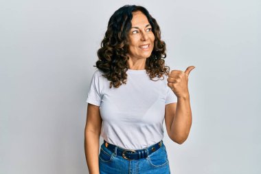 Orta yaşlı İspanyol kadın, günlük beyaz tişört giyiyor, gülümsüyor, gülümsüyor ve başparmağıyla yan tarafı işaret ediyor.. 