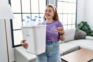 Geri dönüşüm sepetini tutan genç kızıl saçlı bir kadın. Elinde plastik şişelerle arka tarafı işaret ediyor. Baş parmakları havada, kendinden emin gülümsüyor. 