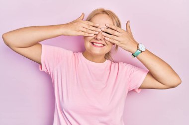 Sıradan pembe tişörtler giyen genç sarışın kadın. Gözleri kapayan, neşeli ve komik gülümseyen eller. kör kavram. 