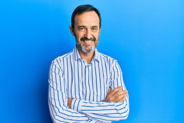 Orta yaşlı İspanyol bir adam sıradan kıyafetler giyip kollarını kavuşturup kameraya bakarken gülümsüyor. pozitif kişi. 