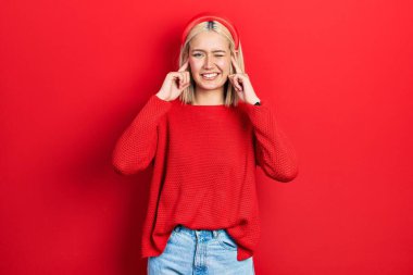 Gürültülü müzik sesi için kulaklarını parmaklarıyla kapatan gündelik kırmızı süveter giyen güzel sarışın kadın. sağır kavramı. 