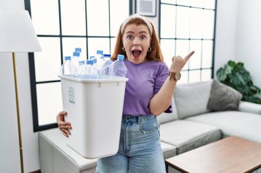 Elinde geri dönüşüm sepetini tutan genç, kızıl saçlı bir kadın. Plastik şişelerle, parmakları yana dönük, ağzı açık, şaşkın bir ifadeyle şaşırdı.. 
