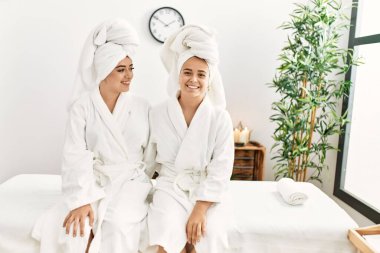 Bornoz giymiş bir çift birbirlerine sarılıyorlar güzellik merkezinde masaj masasında oturuyorlar.