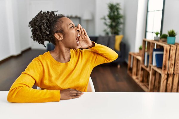 Young african american woman wearing casual clothes sitting on the table at home shouting and screaming loud to side with hand on mouth. communication concept. 