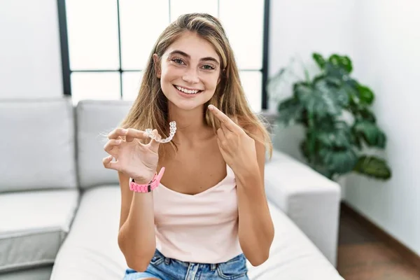 Young blonde woman holding invisible aligner orthodontic smiling cheerful showing and pointing with fingers teeth and mouth. dental health concept.