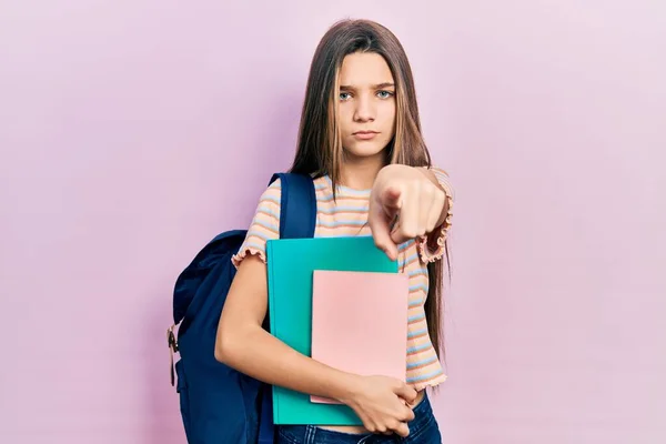 年轻的黑发女孩拿着学生的背包和书本 用手指着相机和你 自信地摆出一副严肃的姿势 — 图库照片