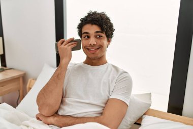 Genç İspanyol adam akıllı telefonda konuşuyor. Yatak odasında yatakta oturuyor..