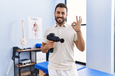 Fizyoterapi merkezinde terapi tabancası tutan yakışıklı İspanyol adam parmaklarıyla imza atıyor, gülümseyerek mükemmel bir işaret veriyor. 