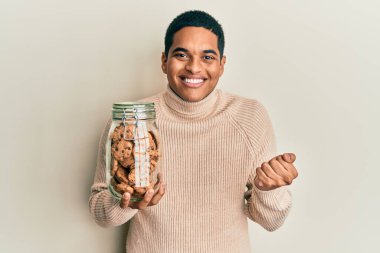 Genç, yakışıklı İspanyol adam elinde çikolata parçacıklı kurabiye kavanozuyla gururla bağırıyor, zaferi kutluyor ve başarıyı kollarını kaldırarak kutluyor. 