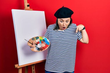 Orta yaşlı İspanyol kadın paletle resim çiziyor. Resim sehpası, parmaklarıyla işaret ediyor. Reklam veriyor, şaşırıyor ve ağzını açıyor. 