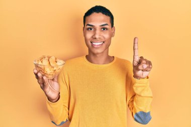 Genç Afro-Amerikalı adam elinde patates cipsi ile gülümsüyor bir fikir veya soru işareti ile mutlu bir yüzle, bir numara 
