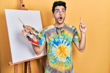 İspanyol asıllı genç adam paletle resim çiziyor. Şaşkın şaşkın duruyor ve parmaklarıyla işaret ediyor ve kollarını kaldırıyor.. 