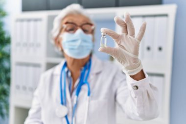 Doktor üniforması giyen yaşlı gri saçlı kadın ve klinikte aşıyı elinde tutan tıbbi maske.