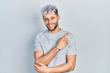 Modern boyalı saçları olan, yüzünde kocaman bir gülümseme olan gri tişörtlü, elleri ve parmaklarıyla kameraya bakan genç İspanyol bir adam.. 