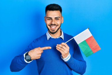 Sakallı genç İspanyol adam, elinde madagaskar bayrağı, gülümsüyor. 