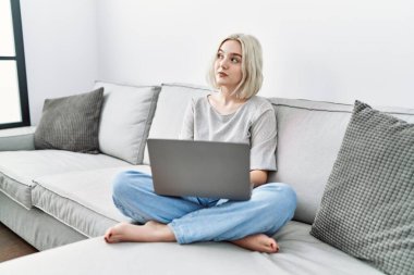 Genç beyaz kadın evinde dizüstü bilgisayar kullanıyor kanepeye oturmuş gülümsüyor ve yan tarafa bakıyor ve düşünüyor.. 