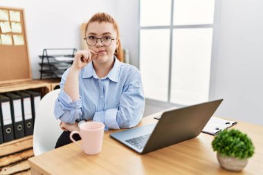 Ofiste çalışan genç kızıl saçlı bir kadın dizüstü bilgisayarın ağzını kullanıyor ve dudaklarını parmaklarıyla kapatıyor. gizli ve sessiz, tabu konuşması 