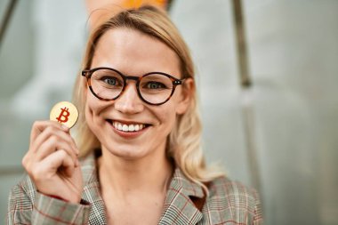 Genç sarışın iş kadını gülümseyerek şehirde Bitcoin tutuyor..