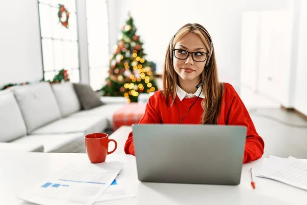 年轻的高加索女孩坐在桌子上 用笔记本电脑在圣诞树旁工作 面带微笑地看着旁边 目不转睛地想着 — 图库照片