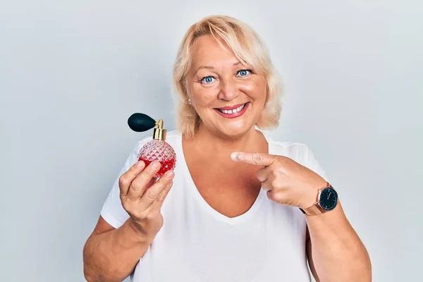 Middle age blonde woman holding perfume smiling happy pointing with hand and finger