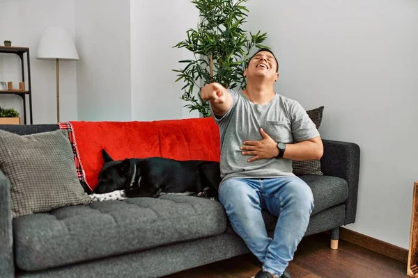 年轻的拉丁男人和狗坐在家里的沙发上嘲笑你 用手指着摄像机 满脸羞愧的表情 — 图库照片