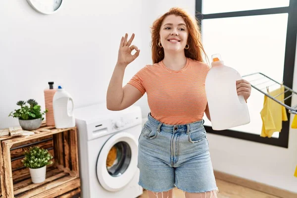 年轻的红头发女人拿着洗涤剂瓶子洗衣服 用手指做手势 微笑友善地表示出极好的象征 — 图库照片