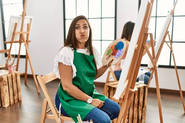 若いヒスパニック系のアーティストの女性が面白い顔で頬をむいてアートスタジオでキャンバスに絵を描いています 口は空気で膨らみ狂気の表情で — ストック写真