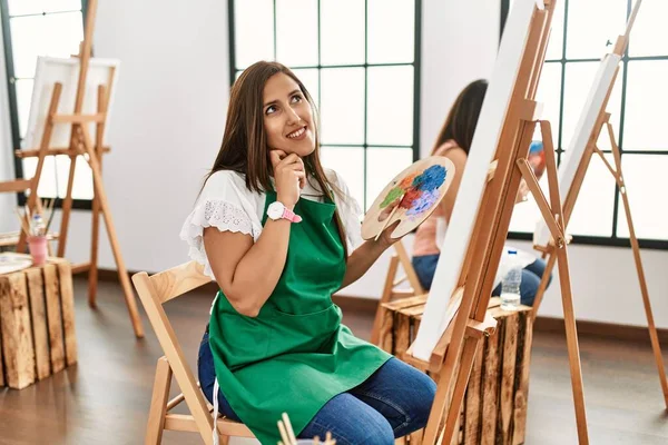 若いヒスパニック系のアーティストの女性は 集中的な表現について考えて顎を手でアートスタジオでキャンバスに絵を描く 思慮深い顔で笑顔 疑わしい概念 — ストック写真