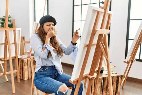 Young hispanic artist woman painting on canvas at art studio smelling something stinky and disgusting, intolerable smell, holding breath with fingers on nose. bad smell