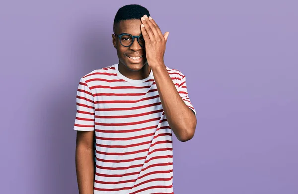 若いアフリカ系アメリカ人の男性は カジュアルなTシャツと眼鏡を手で一つの目を覆い 顔に自信を持って笑顔と驚きの感情 — ストック写真