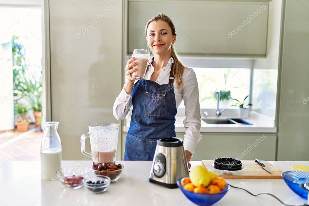 Young blonde woman smiling confident holding smoothie glass at kitchen