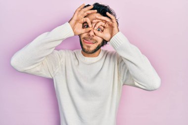 Yakışıklı, beyaz süveter giyen, dürbün gibi jest yapan, gözleri parmaklarından bakan İspanyol bir adam. çılgın ifade. 