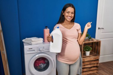 Çamaşır yıkayan hamile bir kadın deterjan şişesini tutuyor. Gülümsüyor. 